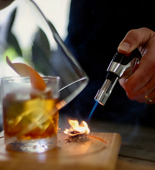 A photo of gifts for couples with a hand using a blow torch on a pile of cedar chips to create smoke for the cocktail next to it.