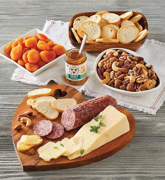 A photo of gifts for couples with a heart shaped cutting board holding cheese and meat surrounded by bowls of mixed nuts and bread and a jar of jam.