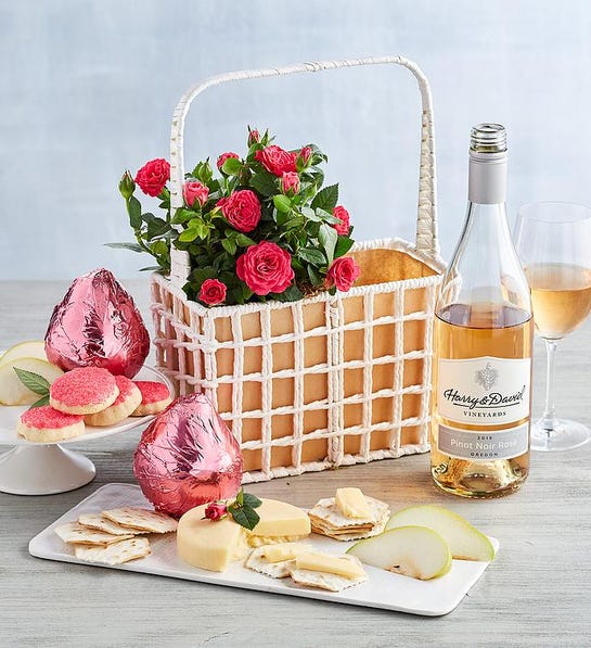 A photo of gifts for couples with a basket of roses next to a bottle of rose, two pears wrapped in pink foil, and a plate of cookies and a plate of cheese, crackers and sliced pears.
