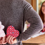 A photo of relationship length with a woman in the background smiling at someone whose back is in the foreground and their holding a stuffed animal and a heart shaped box behind their back.