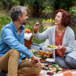 A photo of romantic food with a man and a woman having a picnic outside and feeding each other grapes.