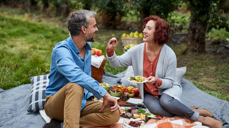 A photo of romantic food with a man and a woman having a picnic outside and feeding each other grapes.