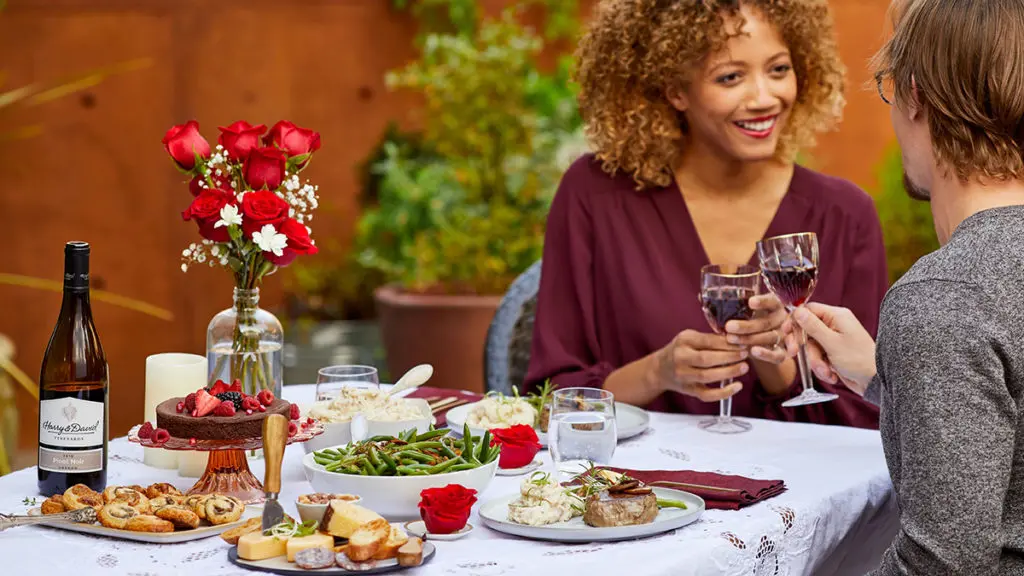 A photo of romantic foods with a couple sitting at a table full of a gourmet meal, wine, and flowers while the two people clink glasses.