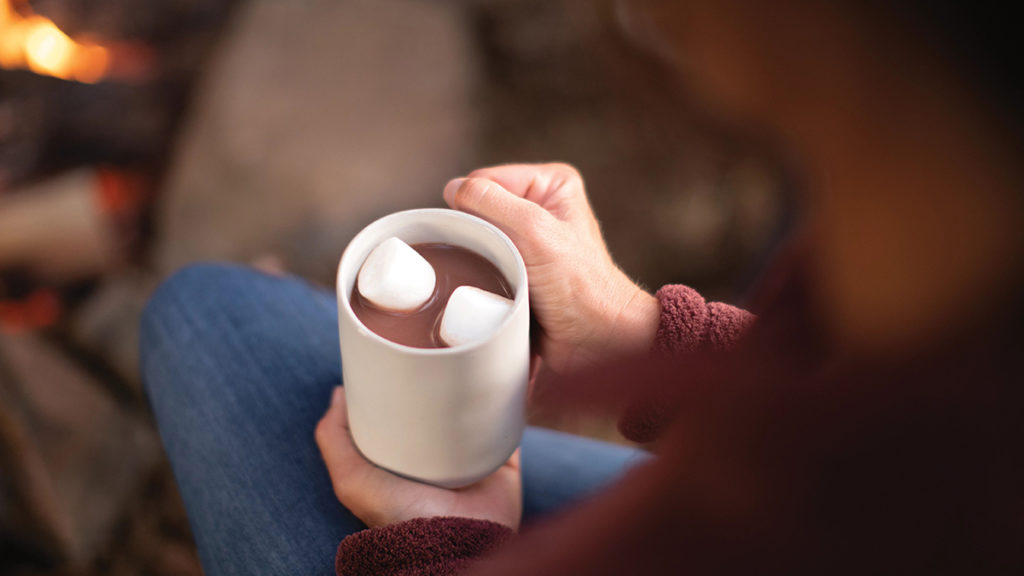 A photo of winter gifts with a cup of cocoa with two marshmallows in it being held by a pair of hands.