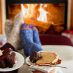 A photo of winter gifts with two pairs of feet resting on a stool with a fire in the background and two plates of cake in the foreground.
