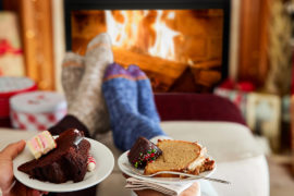 A photo of winter gifts with two pairs of feet resting on a stool with a fire in the background and two plates of cake in the foreground.