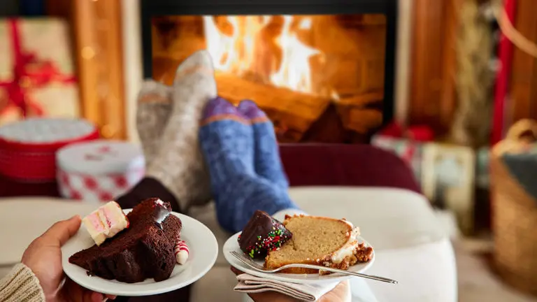 A photo of winter gifts with two pairs of feet resting on a stool with a fire in the background and two plates of cake in the foreground.