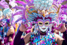 A photo of Mardi Gras with a person in Mardi Gras garb wearing a mask with feathers walking in a parade.