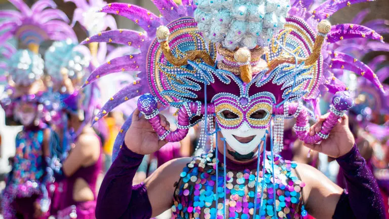 A photo of Mardi Gras with a person in Mardi Gras garb wearing a mask with feathers walking in a parade.