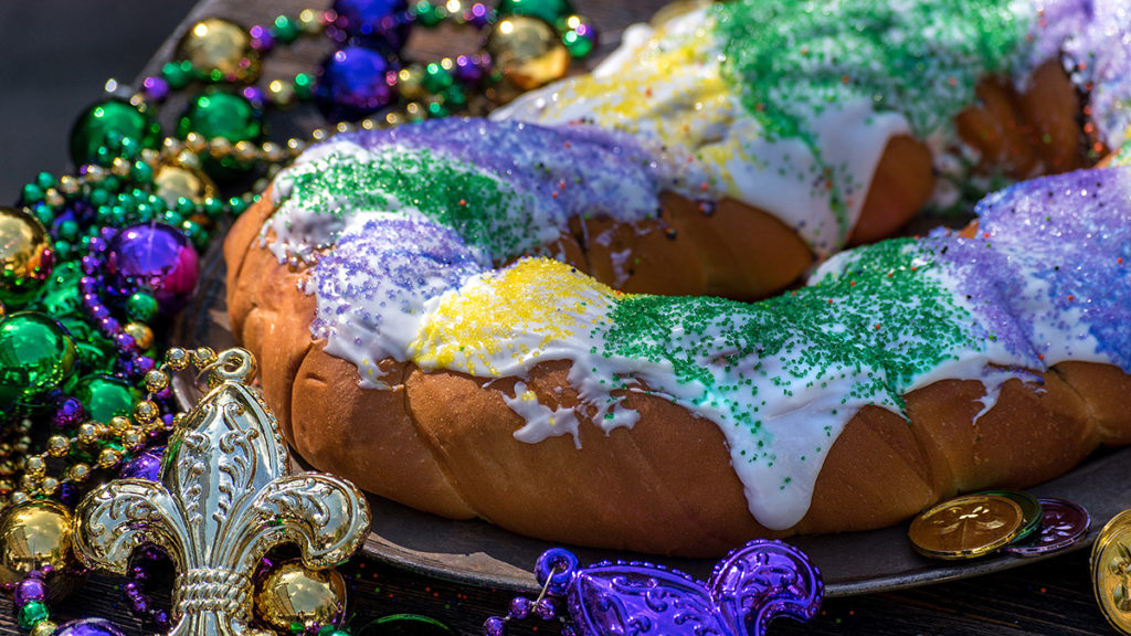 A photo of Mardi Gras with a King Cake covered in frosting and sprinkles with Mardi Gras beads surrounding it.