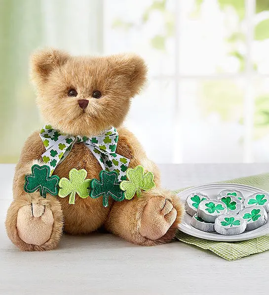 A photo of St. Patrick's Day with a stuffed bear holding a garland of shamrocks sitting next to a plate of chocolates wrapped in tinfoil.