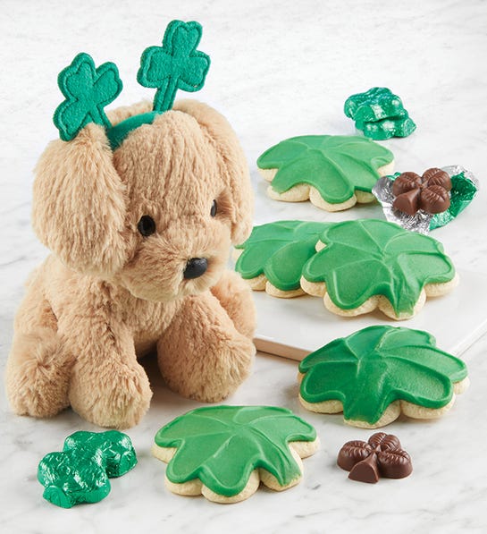 A photo of St. Patrick's Day with a stuffed dog wearing a headband that has two shamrocks on it surrounded by shamrock shaped cookies chocolate.