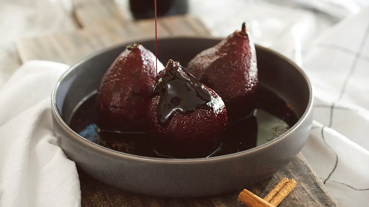 A photo of birthday charcuterie board with three red wine poached pears in a bowl being drizzled with syrup