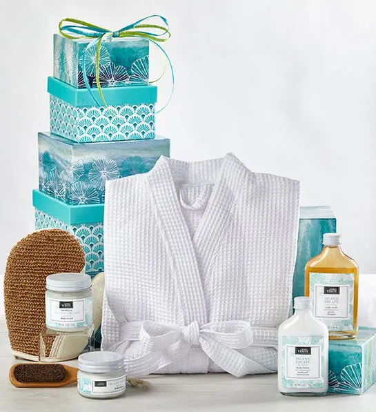 A photo of gifts for women with a stack of blue boxes behind a folded bathrobe and several bottles of lotion and other spa amenities.