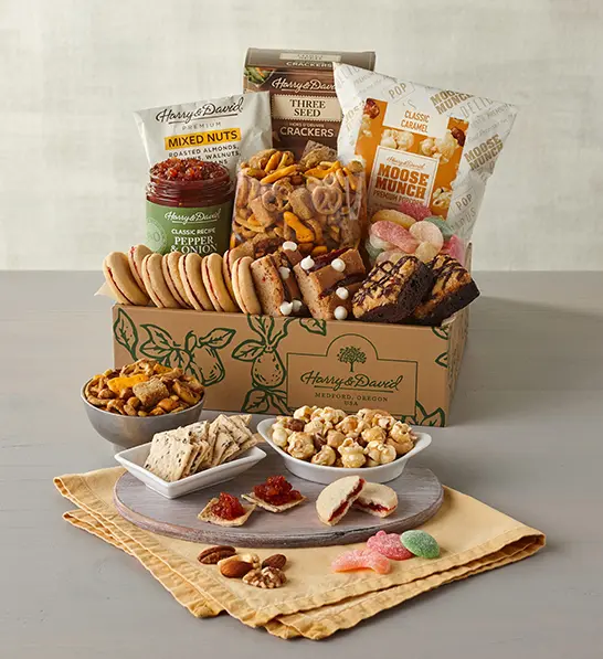 A photo of gifts for women with a box full of sweet and salty snacks with the same items on several plates and bowls in front of the box.