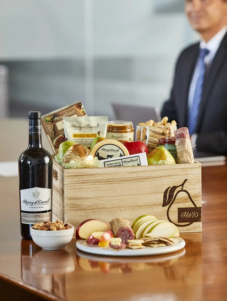 A photo of employee appreciation day with a gift box full of food and snacks on a conference table next to a bottle of wine with a man in a suit in the background.
