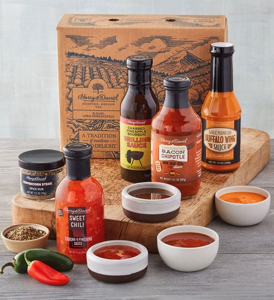 A photo of gifts for men with four jars of grilling sauces on a wooden cutting board with four ramekins full of the same sauces next to the them and two other ramekins full of spices.