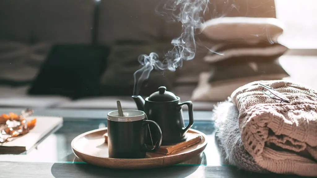 A photo of how to make tea with a pot and a cup on a plate resting on a coffee table next to a pile of blankets