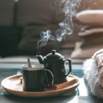 A photo of how to make tea with a pot and a cup on a plate resting on a coffee table next to a pile of blankets