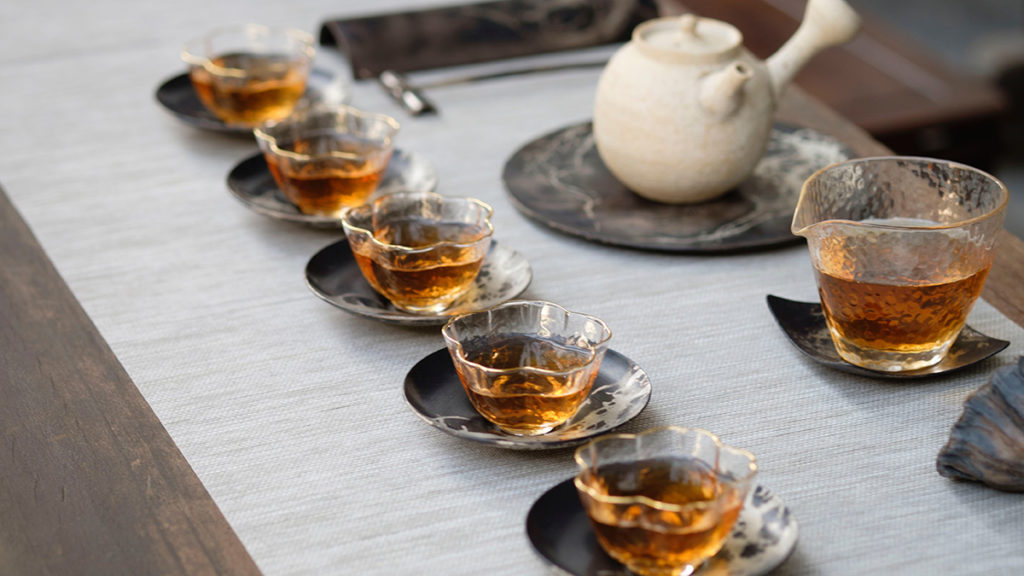 A photo of how to make tea with a row five glass tea cups full of tea and a larger pitcher of tea and a clay pot behind the cups.