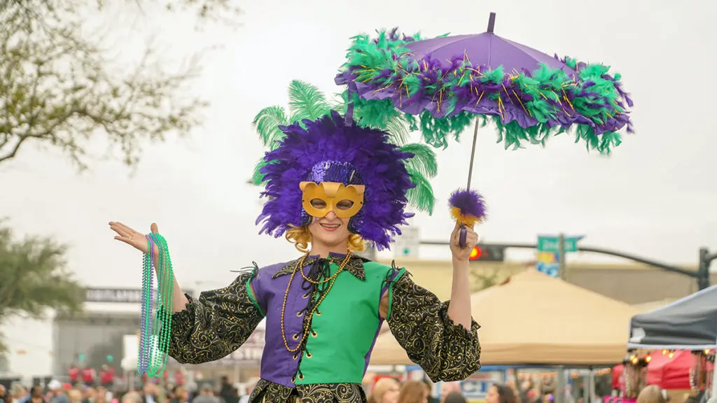 A photo of Mardi Gras with a woman in Mardi dress holding a parasol and beads.