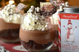 A photo of peppermint cheesecake in two trifle glasses next to a tin of peppermint hot chocolate