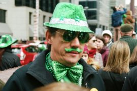 A photo of St. Patrick's Day celebrations with a man wearing a green sparkly fedora and a matching bowtie and a green mustache.