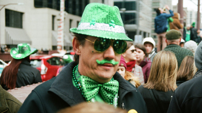 A photo of St. Patrick's Day celebrations with a man wearing a green sparkly fedora and a matching bowtie and a green mustache.