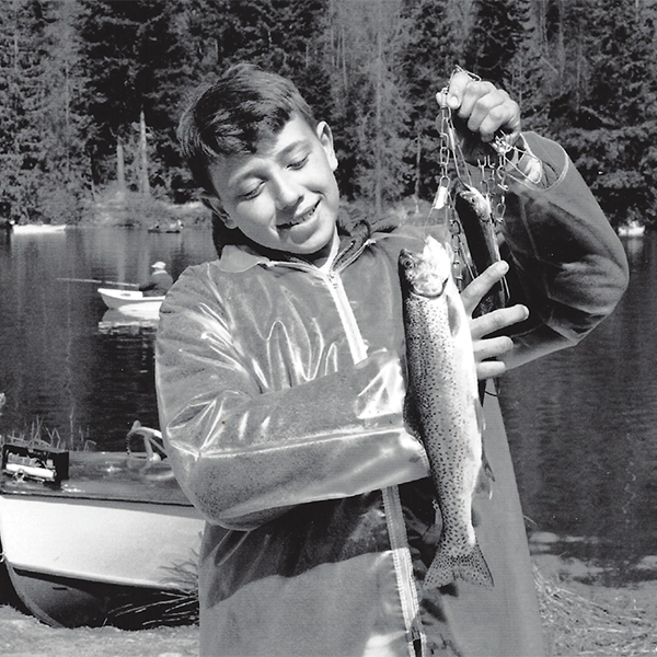 A photo of vital choice with a young boy holding up a fish on a line that's caught in the lake behind him.