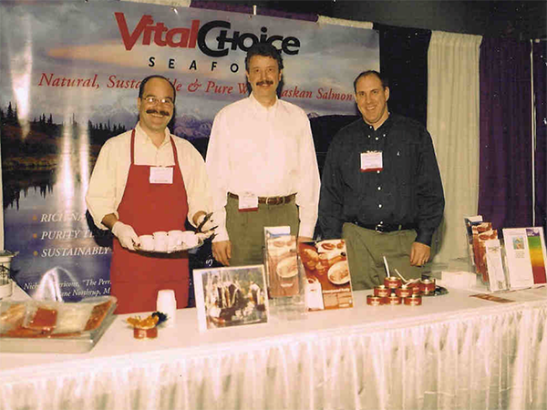 A photo of vital choice with three men standing behind a table and smiling at the camera.