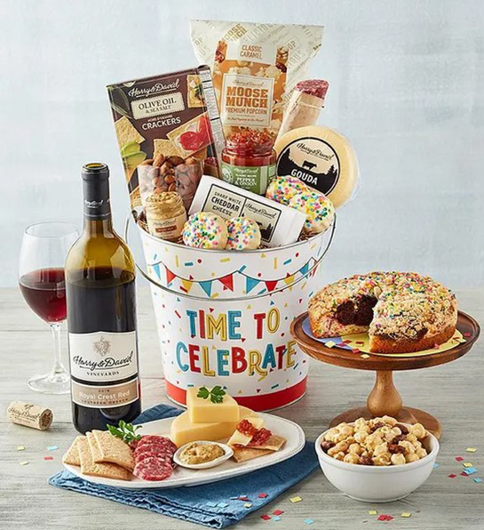 A photo of birthday gifts for sister with a tin that says 'time to celebrate' on it full of charcuterie, cheese and crackers surrounded by the same ingredients and wine.