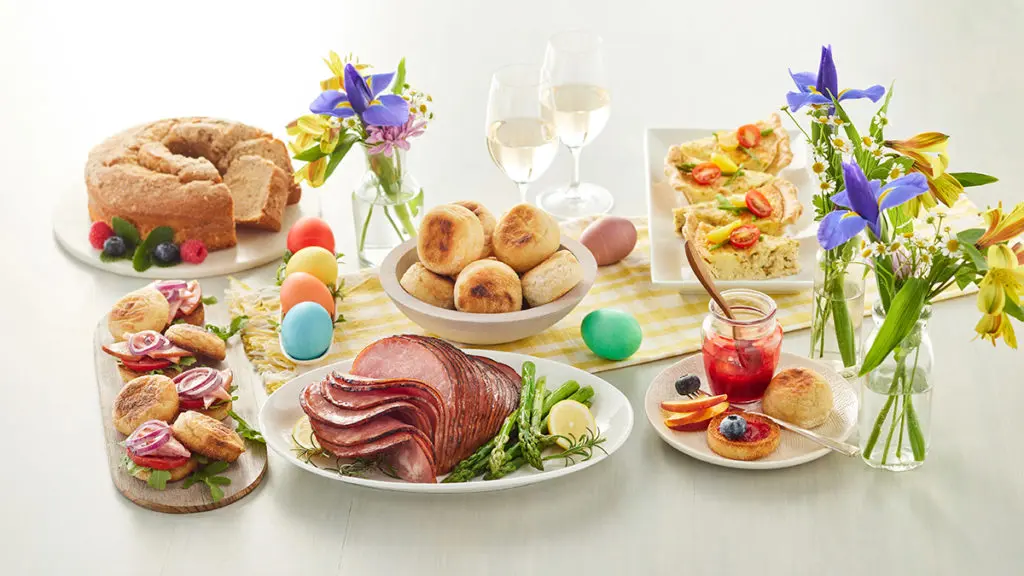 A photo of Easter food with a spread of brunch food like ham, coffee cake, English muffins, and quiche with several bouquets of spring flowers.