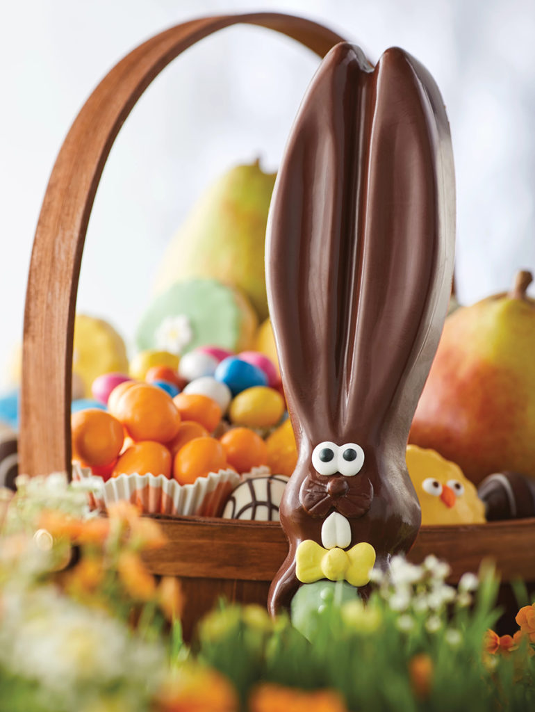 Una foto de comida de Pascua con un conejito de chocolate descansando junto a una canasta de Pascua llena de galletas, frutas y dulces de Pascua.