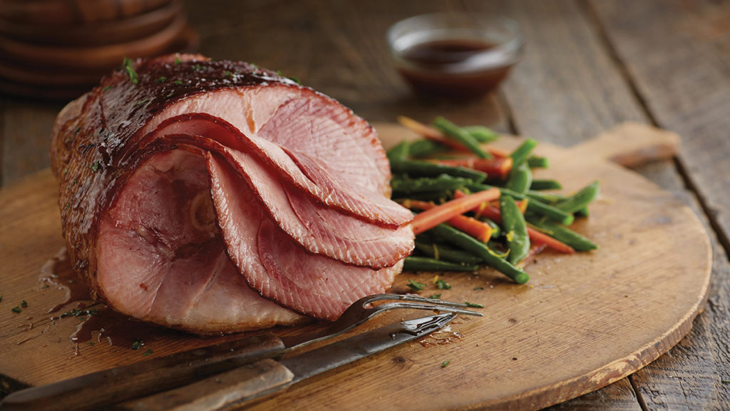 A photo of Easter food with a spiraled ham on a wooden cutting board with a pile of cooked beans and carrots next to it.