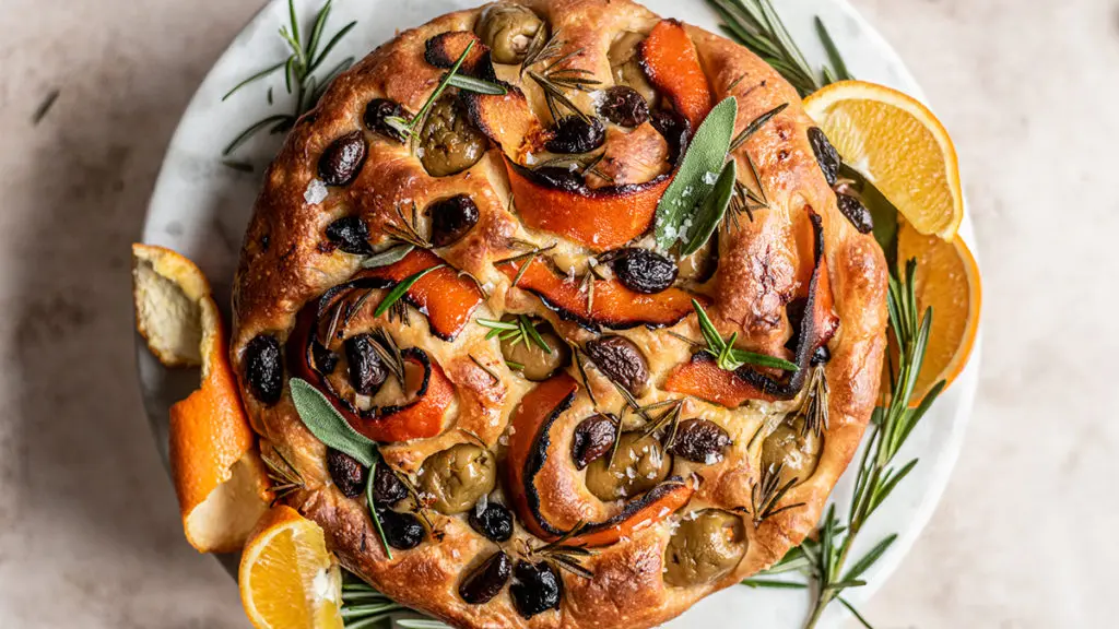 A photo of focaccia with a baked focaccia loaf with olives, citrus peels and rosemary