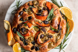 A photo of focaccia with a baked focaccia loaf with olives, citrus peels and rosemary