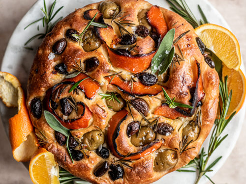A photo of focaccia with a baked focaccia loaf with olives, citrus peels and rosemary