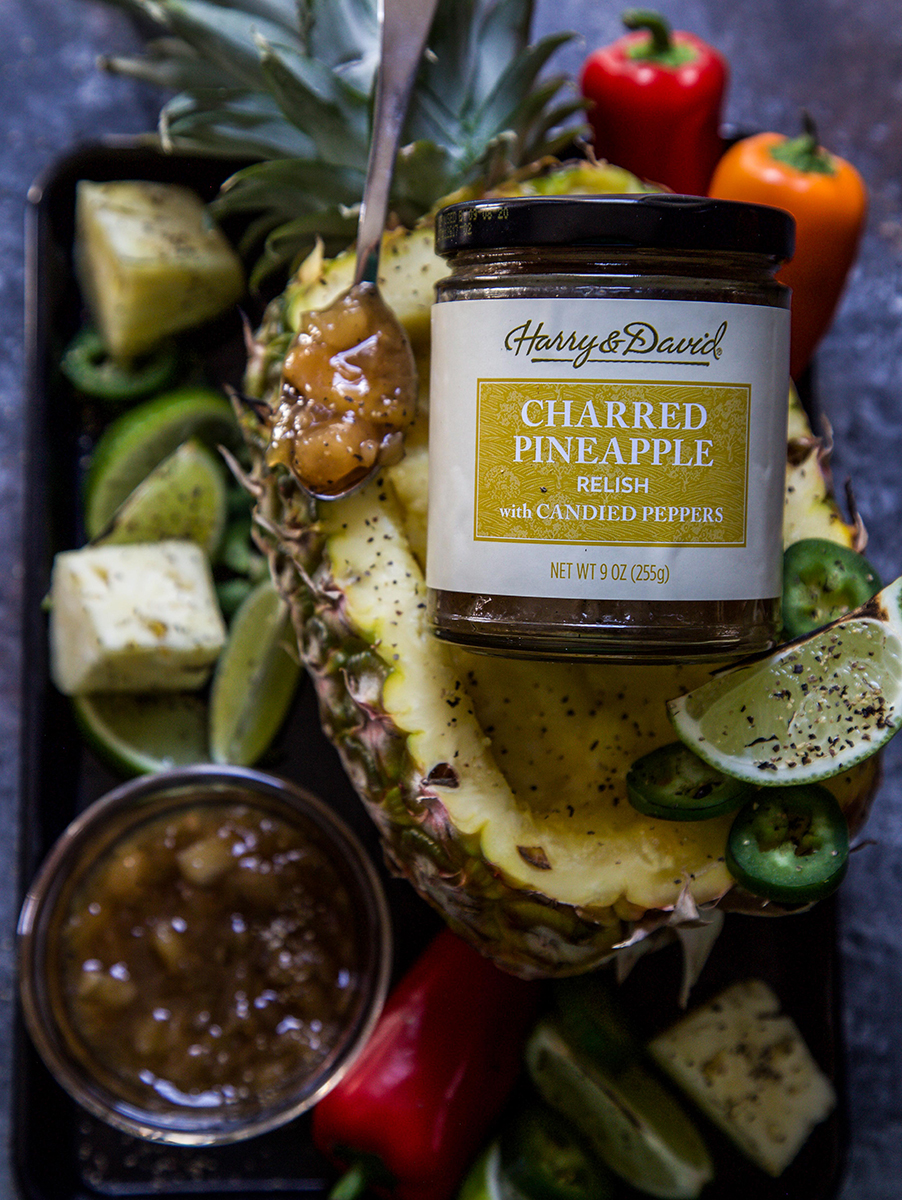 A photo of teriyaki chicken with half a pineapple holding up a jar of Harry & David's charred pineapple salsa surrounded by vegetables and cut limes.