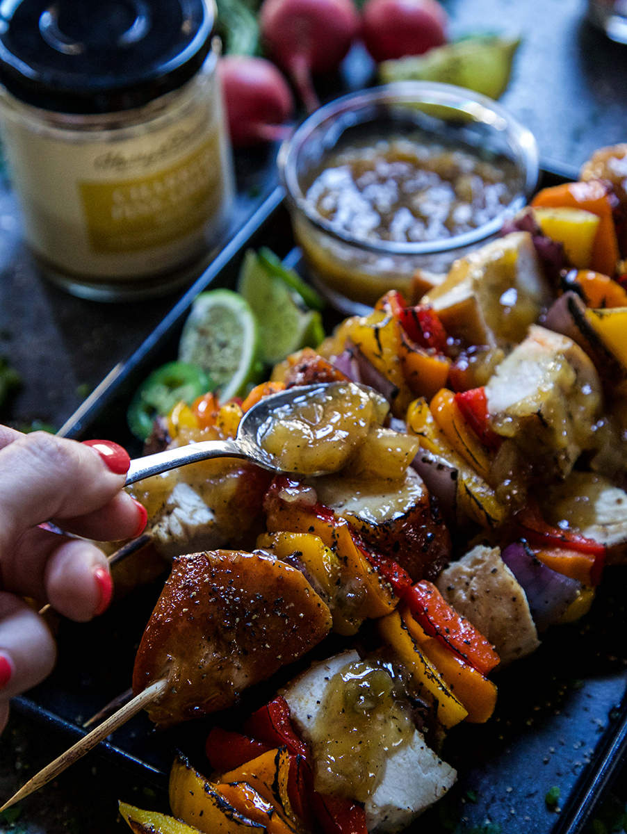A photo of teriyaki chicken with a plate of chicken skewers with a hand drizzling Harry & David's charred pineapple relish over the skewers.