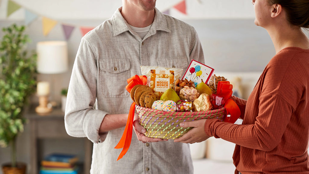 A photo of gifts for hard to buy for with a person handing another person a gift basket full of food