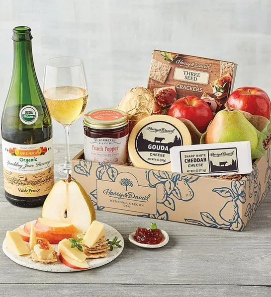A photo of gifts for new moms with a crate of cheese, fruit and crackers next to a bottle of sparkling juice and a plate full of apples and cheese
