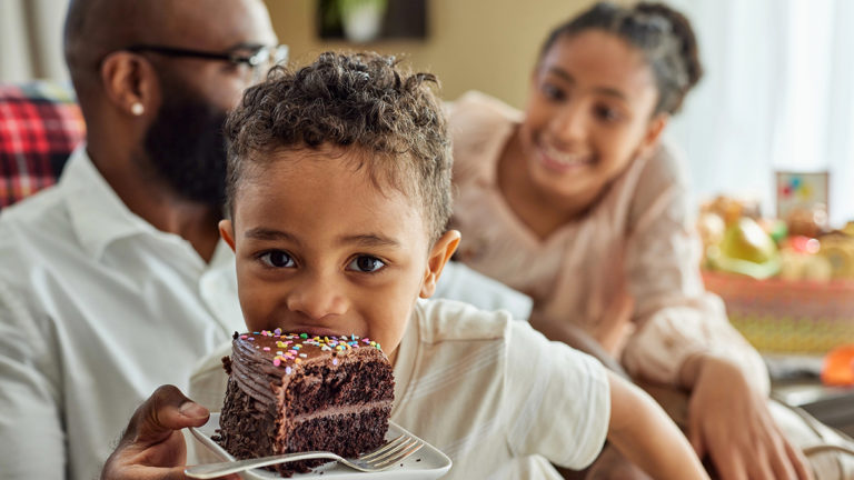 A photo of may birthdays with a boy eating cake with a young girl and older man in the background