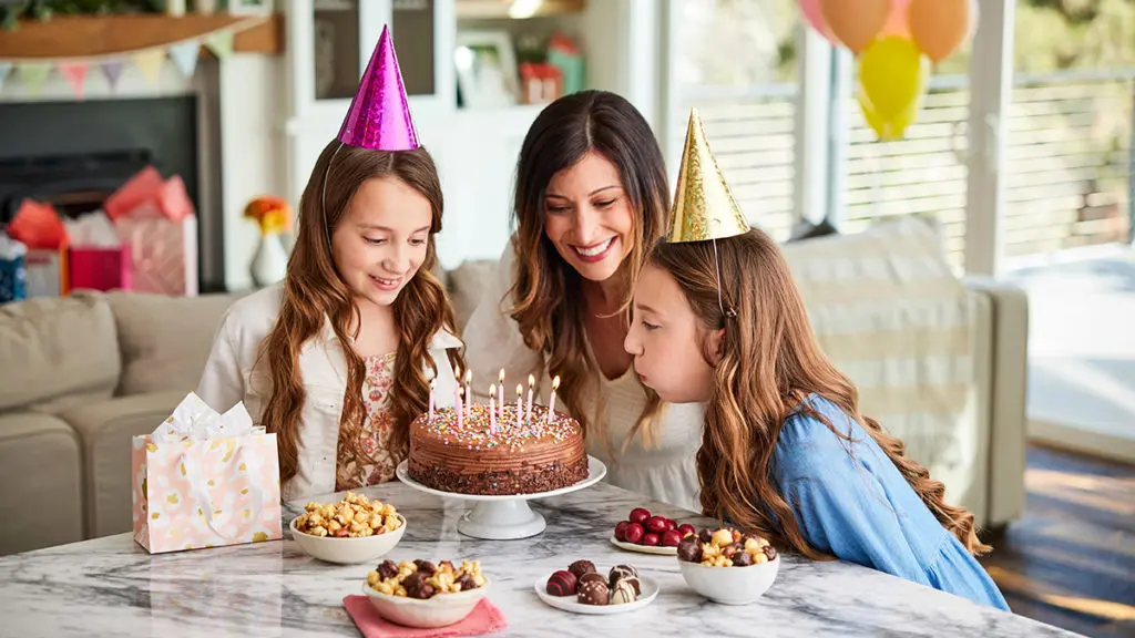 A photo of may birthdays with a pair of twin girls blowing out candles on a cake with their mother watching