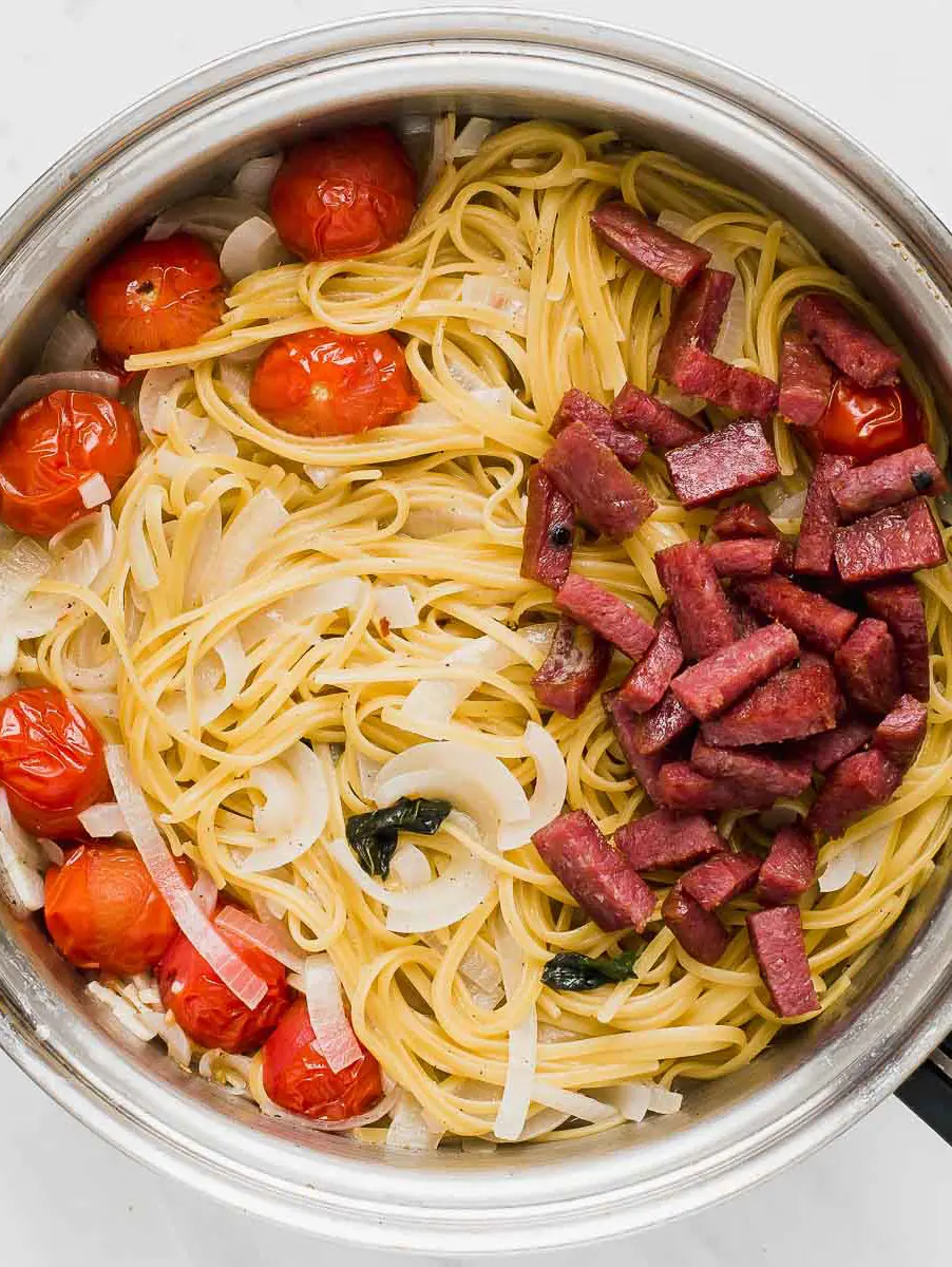 A photo of one pot pasta with a pot full of cooked noodles, tomatoes and salami slices.