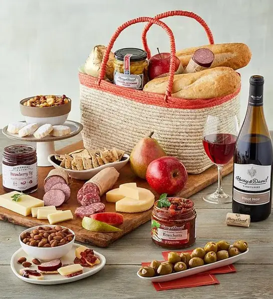 A photo of picnic with a basket full of cheese, bread and other snacks surrounded by the same ingredients and a bottle of wine
