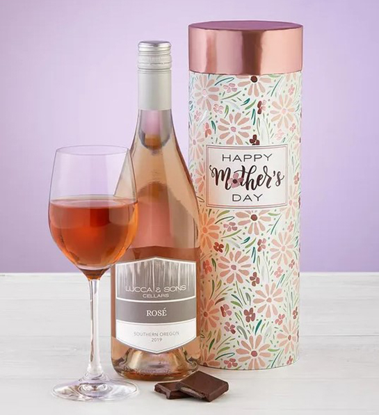 A photo of wine gifts for mom with a bottle of rose wine next to a full glass and a few bits of chocolate and a tube shaped box