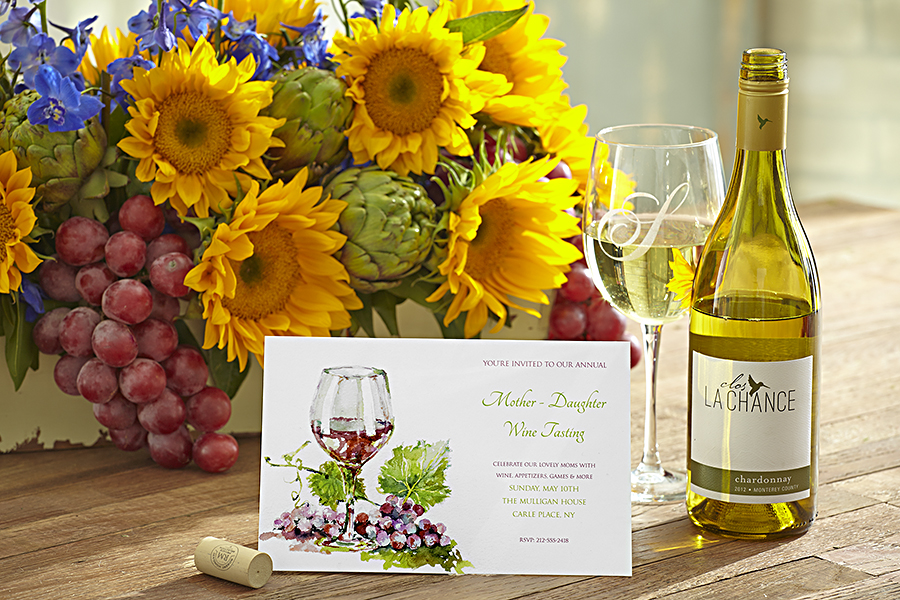 A photo of mother's day wine tasting with a bouquet of flowers behind an invitation to a wine tasting next to a bottle and glass of wine.