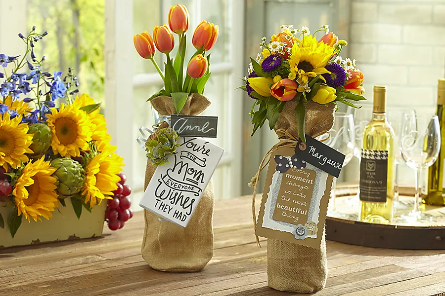 A photo of mother's day wine tasting with two bottles of wine in burlap sacks with flowers coming out of the top.