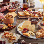 A photo of charcuterie with a table full of meats, cheese, nuts, wine and other snacks