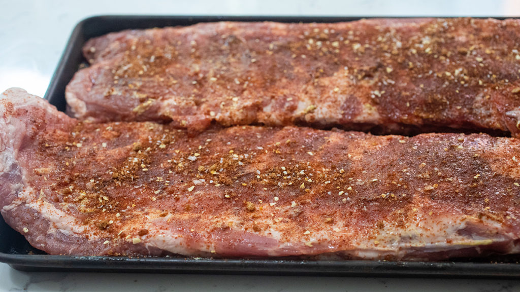 A photo of grill ribs with two racks of ribs on a platter with seasoning rubbed on them